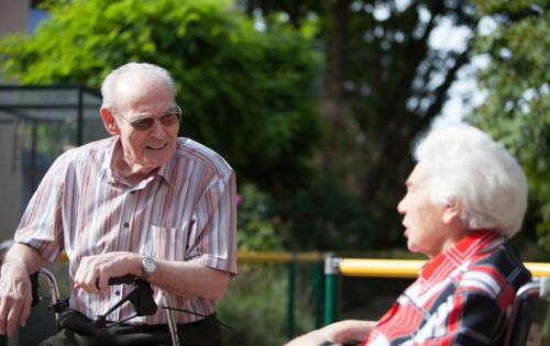 Ein alter Mann und eine alte Frau im Gespräch: Er trägt ein gestreiftes Hemd und stützt sich auf seinen Rollator. Sie sitzt rechts von ihm in einer rot-schwarz-karierten Bluse. Sie sitzen draußen, es ist sonnig.