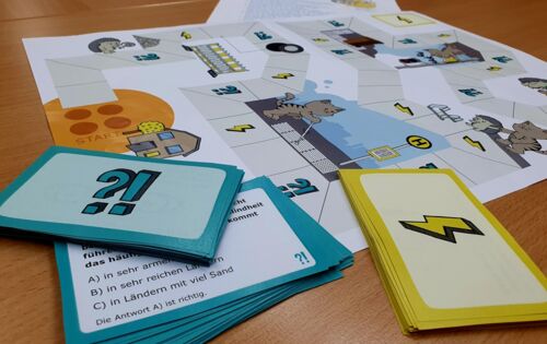 Auf einem Tisch liegen das Spielbrett und zwei Kartenstapel mit unterschiedlichen  Symbolen: auf dem einen ein Frage- und ein Ausrufezeichen, auf dem anderen ein Blitz.