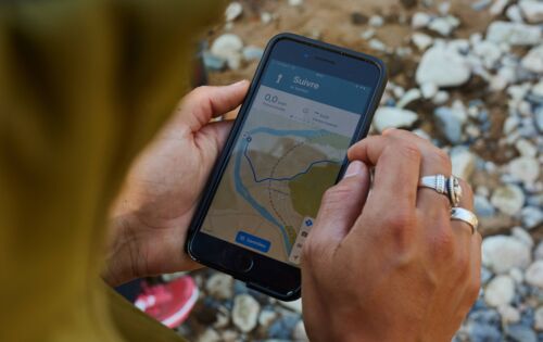 Jemand steht draußen auf Steinen und hält ein Handy mit der Navigations-App Komoot in der Hand. Darauf ist eine Route zu sehen. Der Mensch hat silberne Ringe und trägt eine grüne Jacke.