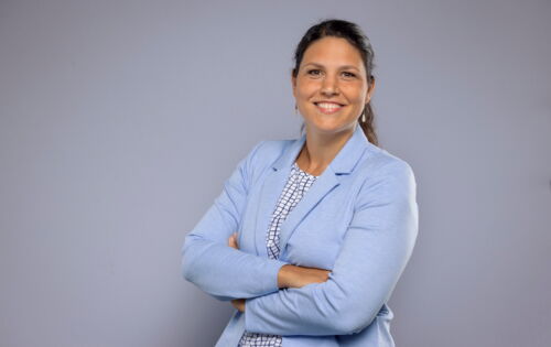 Sarah Karge, eine Frau mit brauen, langen Haaren, steht mit verschränkten Armen vor einem neutralen Hintergrund. Sie trägt einen hellblauen Blazer über einer gemusterten Bluse. Sie lächelt offen in die Kamera.