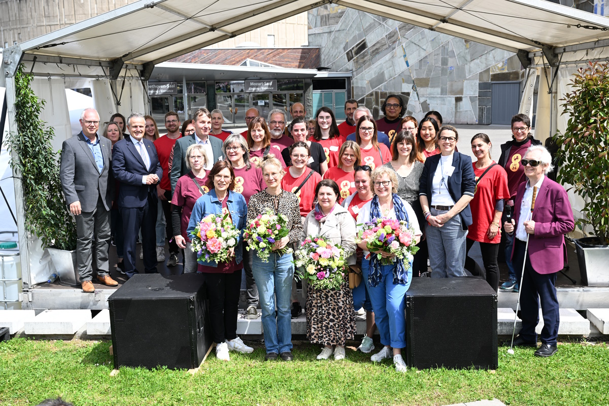 Unter dem weißen Zelt der Außentribüne sammeln sich symbolisch viele Helferinnen und Helfer, die das Louis Braille Festival möglich gemacht haben. Einige tragen das offizielle Festival-T-Shirt, vier von ihnen halten große Blumensträuße.